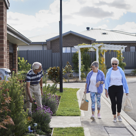 Two women walking down a street at a retirement village and greeting a man watering his front garden