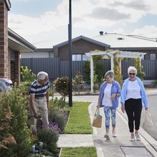 Two women walking down a street at a retirement village and greeting a man watering his front garden