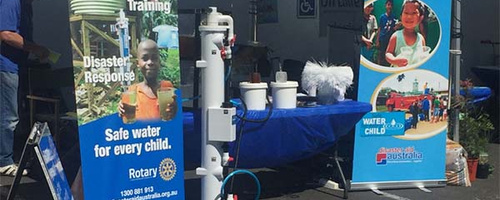 A table with a blue cloth holds water sanitation equipment. A large water sanitation pipe machine stands in front of the table. Rotary’s safe water pull-up banners sit on either side of the table.