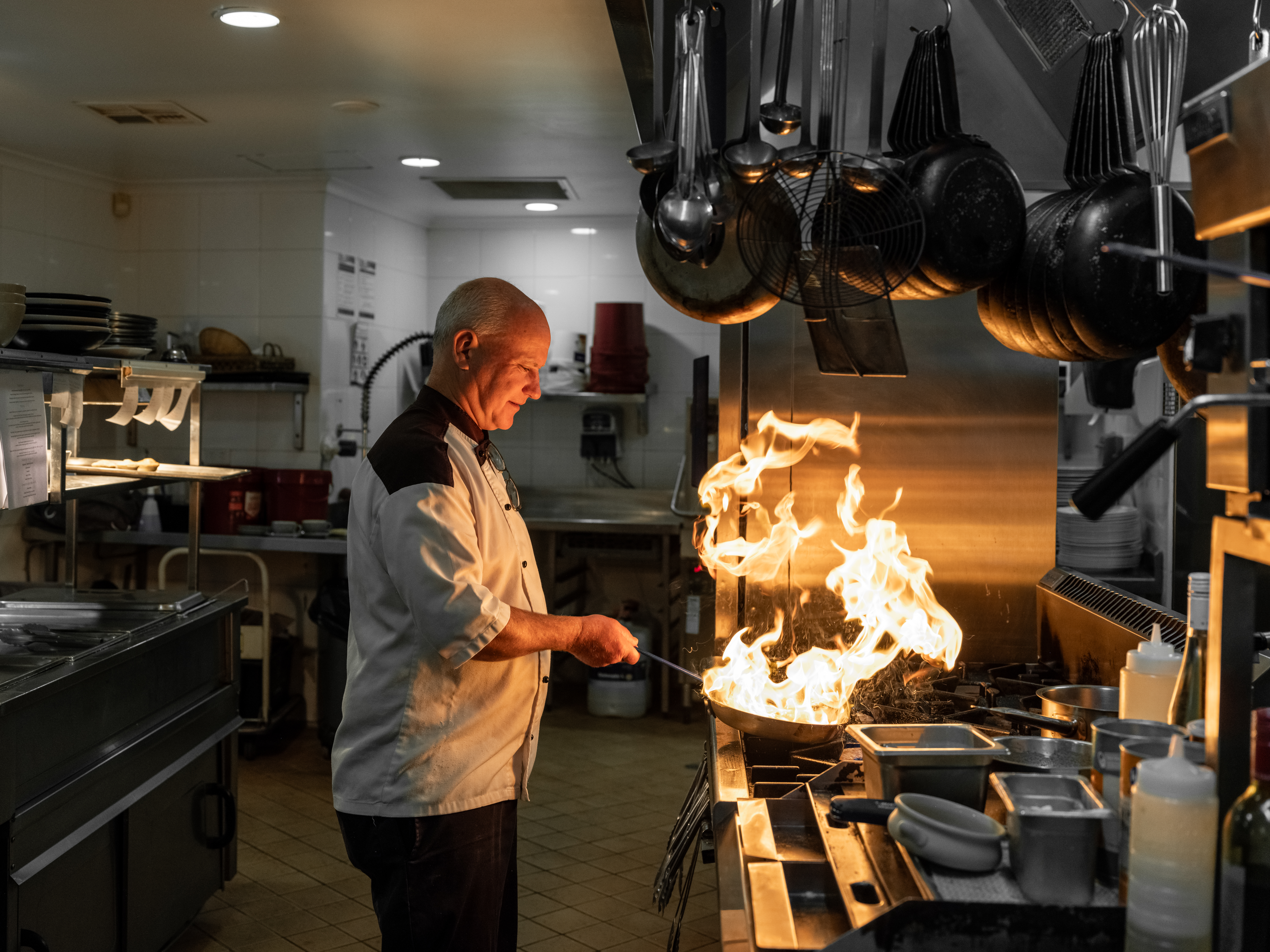 Ardency Aroona chef Colin standing at the stove of the kitchen at Brookside Restaurant and frying something in a pan with fire.