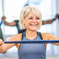 A smiling woman is doing resistance band exercises as part of a gym class for seniors and older adults.
