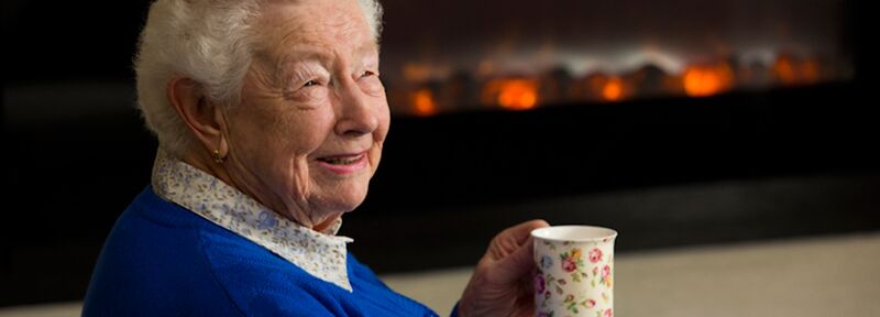Retirement village resident Judith sits by a fireplace drinking from a floral coffee cup. She’s smiling, wearing a royal blue jumper with a blue floral blouse.