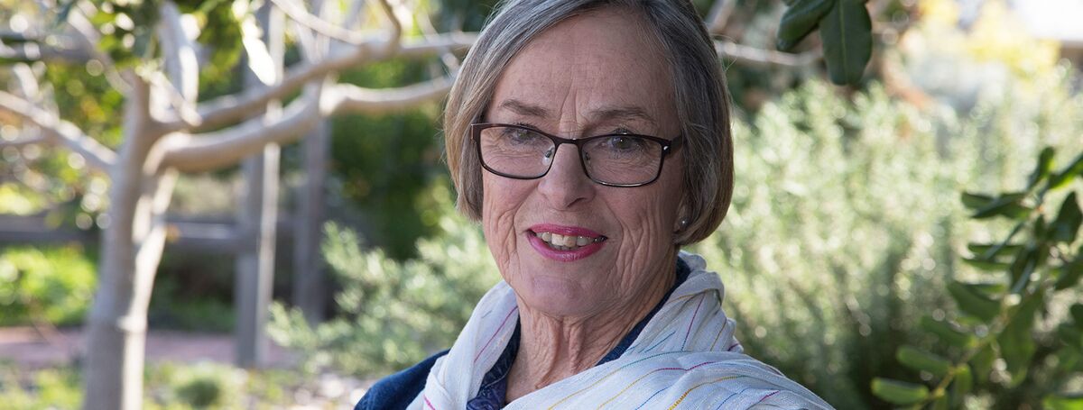 Retirement village resident Rosslyn sits in a garden, looking at the camera, smiling. She has a blonde bob and wears glasses.