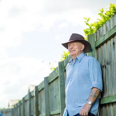 Retirement village resident Don Glasby wears a blue collared shirt and a brown Akubra hat. He’s standing next to a fence on a bright, sunny day.