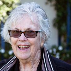 Retirement village resident Betty Mounser smiles at the camera. She has white hair and wears sunglasses while sitting in a garden.