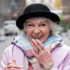 A stylish woman wears a black bowler hat over her grey hair and a pale pink jacket with a blue, white and purple scarf.