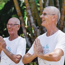 Retirement village residents Michael and Torjborn stand side by side wearing white t-shirts and smiling while holding their hands in a prayer pose. 