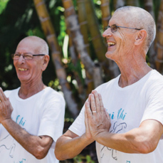 Retirement village residents Michael and Torjborn stand side by side wearing white t-shirts and smiling while holding their hands in a prayer pose. 