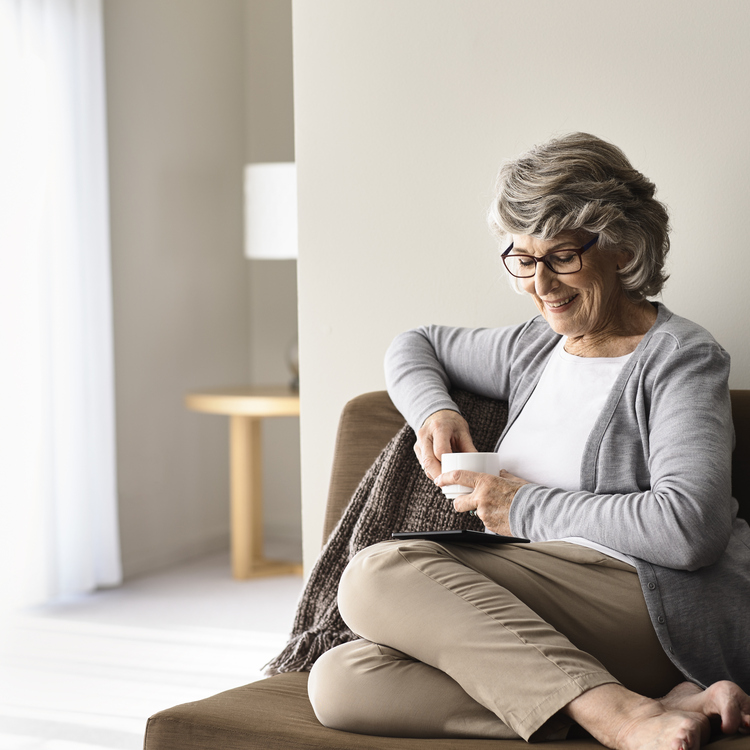 A senior lady sitting on a couch with a cup of tea relaxing and reading with a kindle