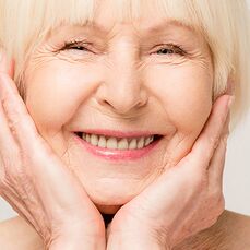 A close-up of an elderly woman’s face. She’s smiling and wearing elegant makeup with pink lipstick, following the best makeup routine for mature skin.