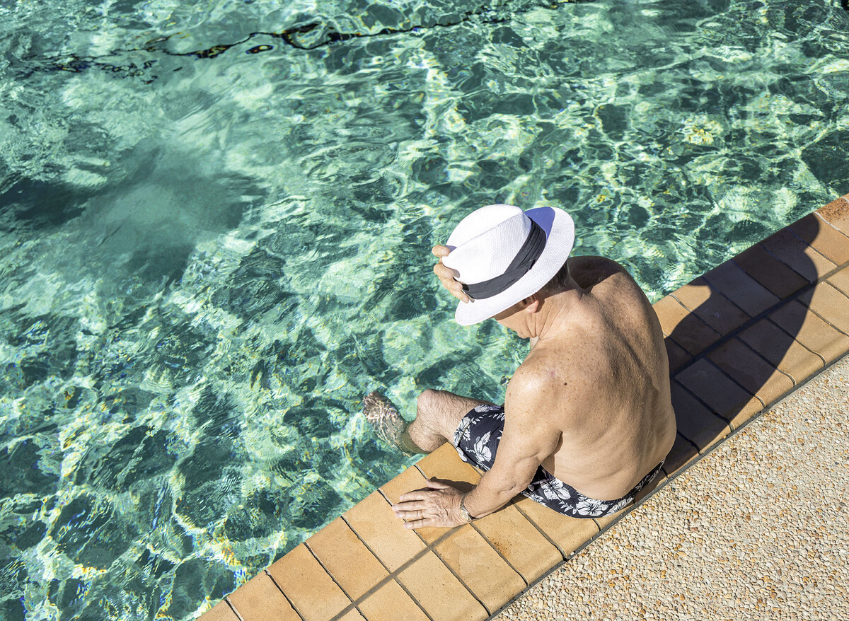 A man wearing a sun hat sitting by a pool with his legs in the water